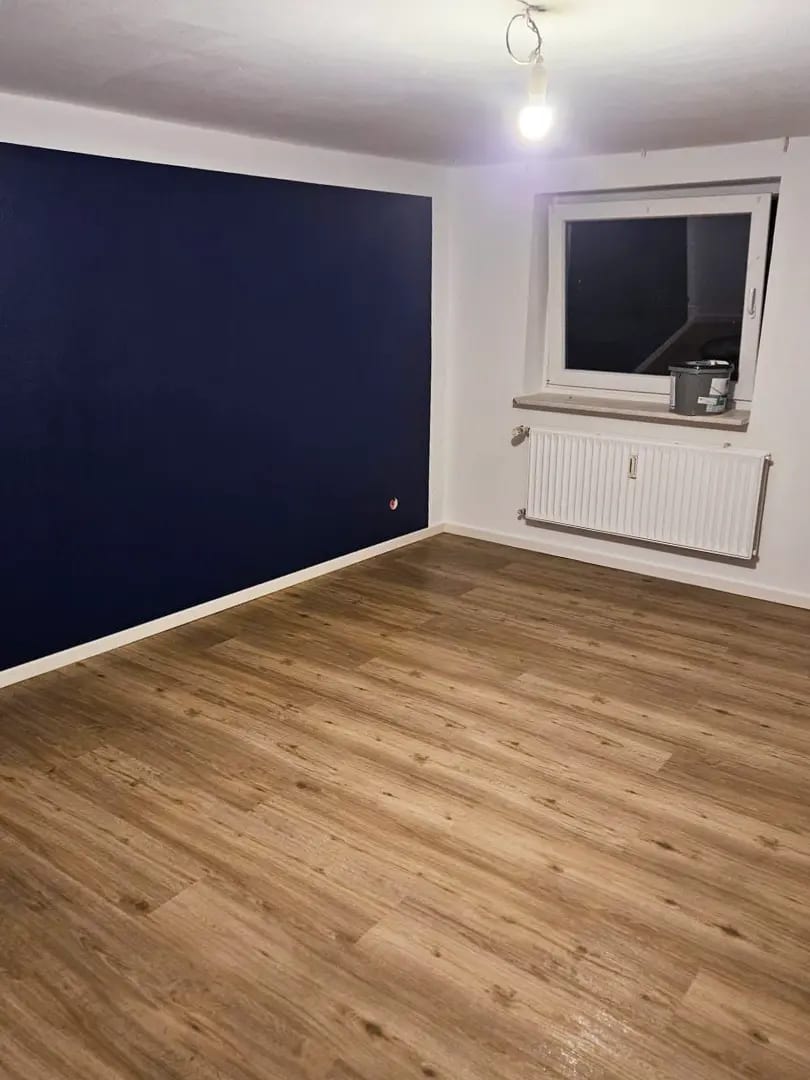 Zimmer mit dunkelblau abgesetzter Wand, Heizkörper unter dem Fenster und neu verlegtem Vinylboden